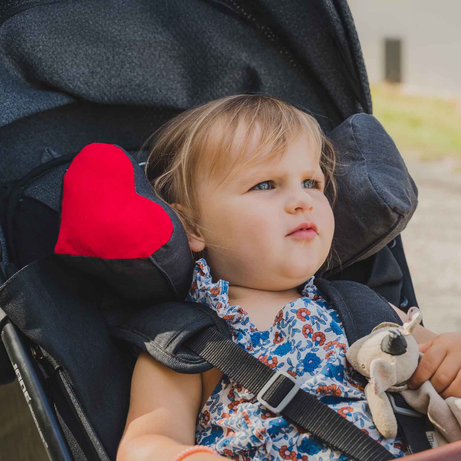 Petite fille dans sa poussette avec coussin Cosy Club pour améliorer le confort de la tête. Coussin gris avec pièce en forme de cœur rouge sur le côté gauche.