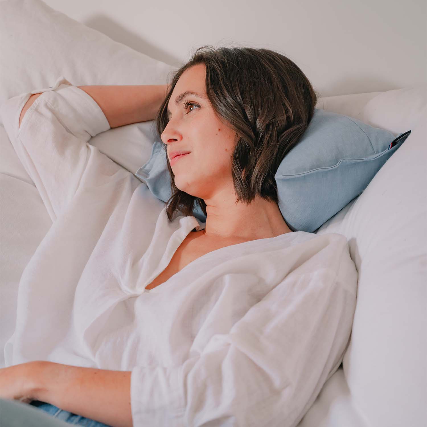Jeune femme paisiblement installée sur son lit, souriante, avec coussin cervical SALVADOR de Cosy Club derrière le cou, coloris bleu pastel. Elle est souriante et semble très détendue. Sensation de confort.