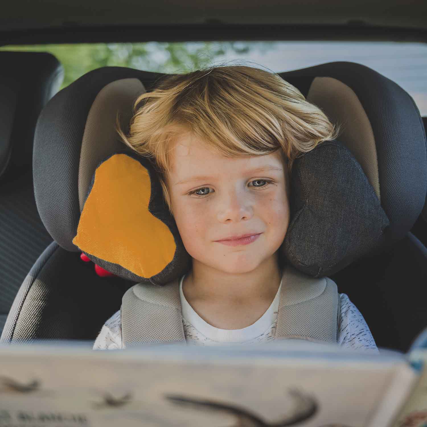 Enfant en train de lire un livre dans son siège auto. Il a la tête bien maintenue grâce au coussin TEODOR de Cosy Club qui comble le vide de chaque côté. Le tissu du coussin est gris avec une pièce en forme de cœur en velours jaune.