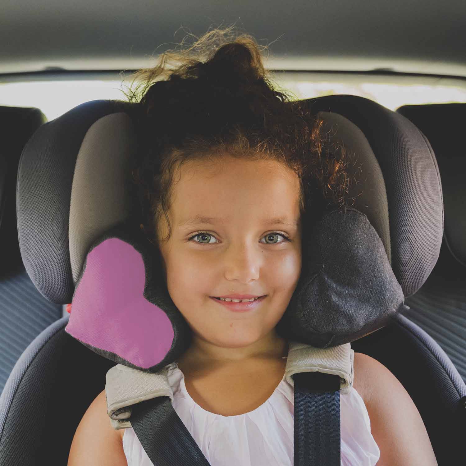 Enfant souriante avec la tête bien calée dans son siège auto avec le coussin TEODOR. Tissu gris anthracite et cœur en velours rose.