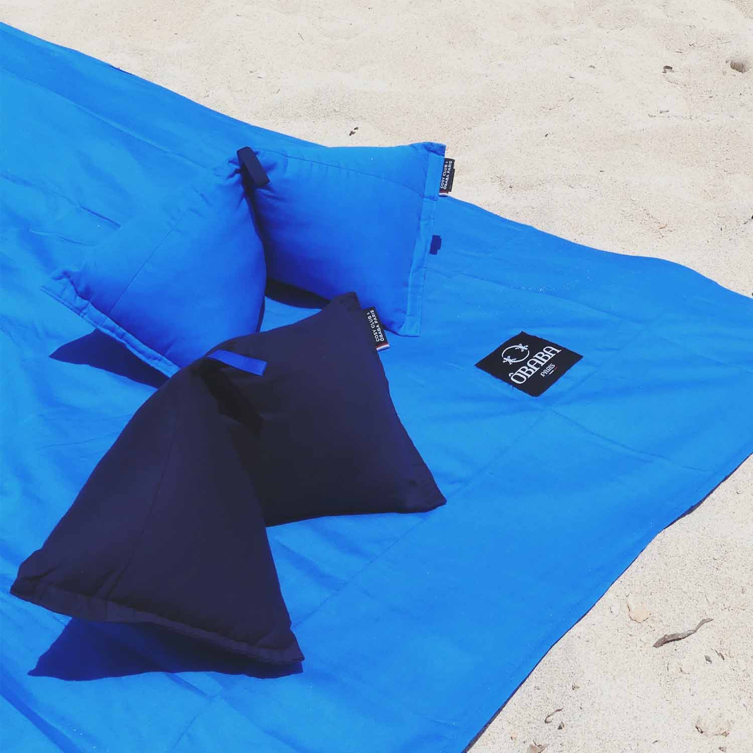 Drap de plage Obaba bleu et deux coussins cervicaux (bleu et bleu marine) disposés sur le sable