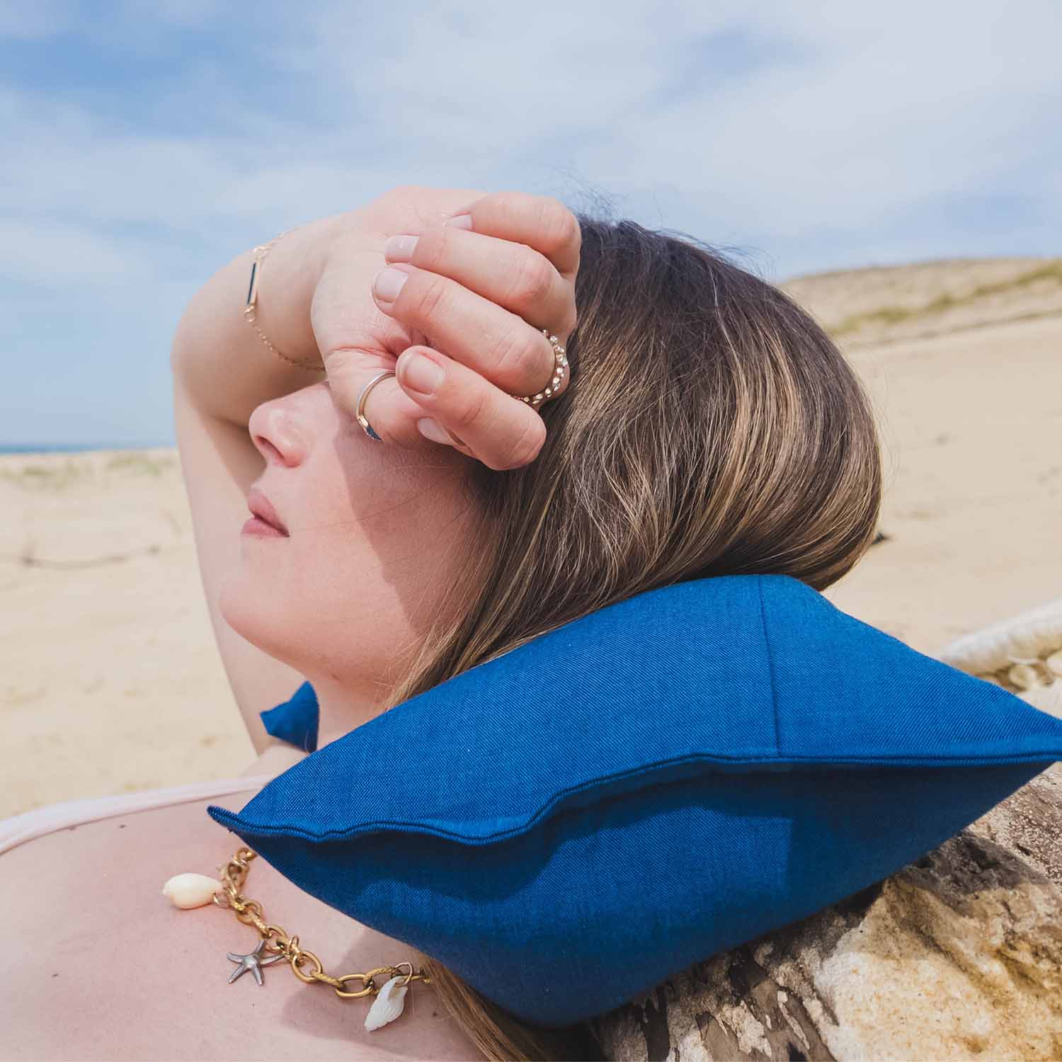 Femme allongée sur le sable, adossée à un bois flotté, avec coussin cervical Cosy Club bleu derrière le cou pour éviter les tensions à la nuque